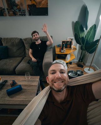 Two men smile in a living room, one taking a selfie in the foreground and one waving from a gray couch in the backgro...