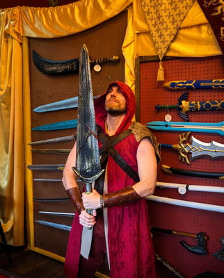 A man in a red hooded costume holds a large decorative sword while posing in front of a pegboard display of replica w...
