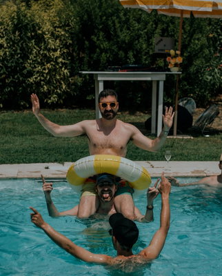 Three men celebrating in an outdoor pool in Tuscany, one sitting on another's shoulders with a yellow float around them.