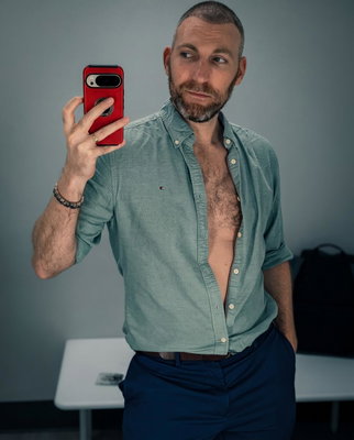 A man takes a mirror selfie holding a red phone wearing an open teal button down shirt and navy trousers in an office...
