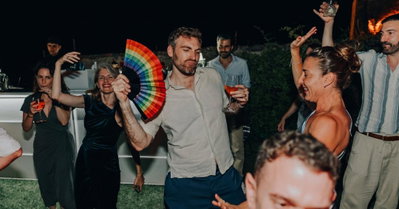 A man dances outdoors at night holding a rainbow fan and a wine glass, surrounded by a group of people at an evening ...