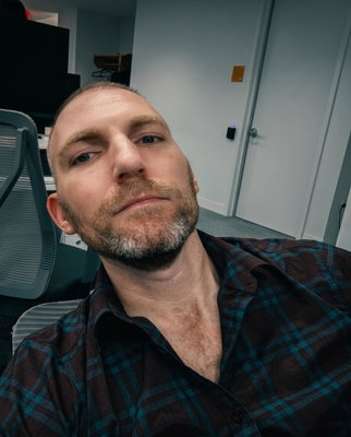 A man with short cropped hair and a beard takes a selfie while seated in an office chair in an indoor workspace.